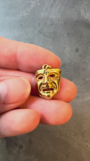 Gold mask pendant turned in hand on a grey background